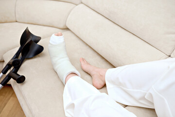 Broken leg in a plaster cast, near a crutches. Youthful woman with a foot bone fracture is taking it easy on the couch. Toe, talus, navicular, cuboid bone fracture. Injury, trauma, recovery concept - obrazy, fototapety, plakaty