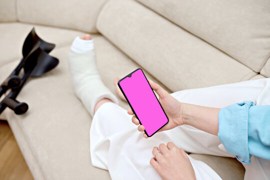 Woman resting on the couch at home with cast on  fractured leg using a phone. Adult checks possible symptoms with professional traumatologist. Concept of online patient counseling and telemedicine.