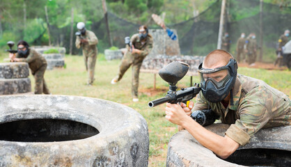 Fototapeta premium Portrait of young man paintball player in mask who is aiming in opponents on paintball field