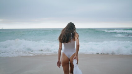 Back view woman walking on stormy ocean waves wearing white sexy swimsuit. 
