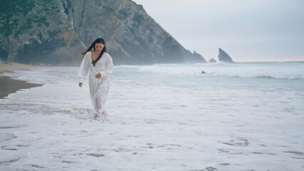 Woman walking sea waves in white cape cloudy evening. Girl enjoy gloomy beach.