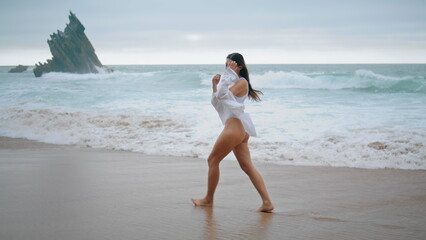 Girl stepping wet sand ocean beach wearing sexy white swimsuit cloudy day. - obrazy, fototapety, plakaty
