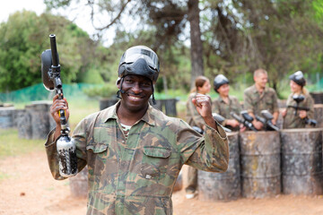 Fototapeta premium Smiling afro american man paintball player in camouflage posing with gun outdoors