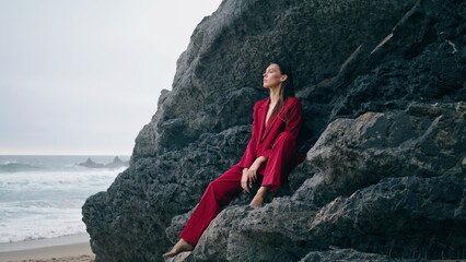 Woman sitting rock ledge in elegant red suit. Stylish girl looking gloomy ocean.