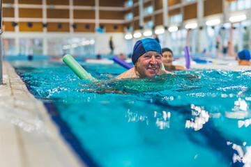 smiling senior Caucasian man with blue swimming cap using a swim noodle in the pool. High quality photo
