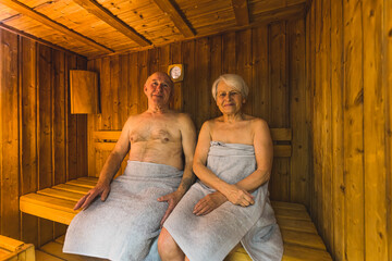 aged Caucasian couple with towels sitting in the wooden sauna and smiling, medium full shot,...