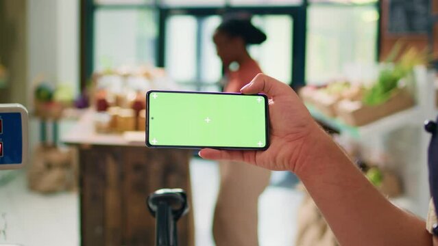 Seller showing greenscreen on phone display, presenting isolated mockup template on gadget in local organic zero waste store. Young man vendor using blank chromakey copyspace screen.