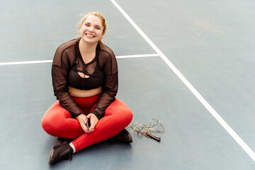 Smiling plus size woman resting after jumping rope, holding smartphone. Sport, workout, technology...