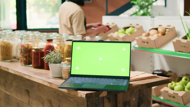Greenscreen laptop in local eco store with crates of organic homegrown produce and bulk products in jars on shelves. PC running isolated display with mockup copyspace template.