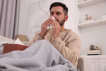 Sick man with tissue blowing nose on sofa at home. Cold symptoms