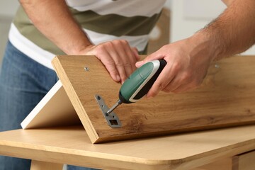 Man with electric screwdriver assembling furniture at table indoors, closeup