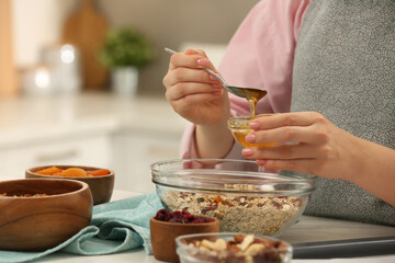 Making granola. Woman adding honey into bowl with mixture of oat flakes and other ingredients at table in kitchen, closeup