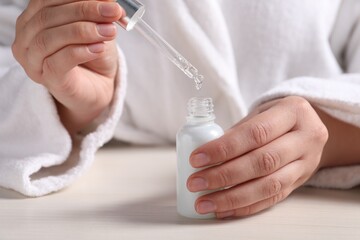 Woman dripping cosmetic serum from pipette into bottle at white table, closeup