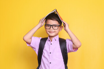 Happy  schoolboy in glasses wearing school bag put a book on his head over yellow background