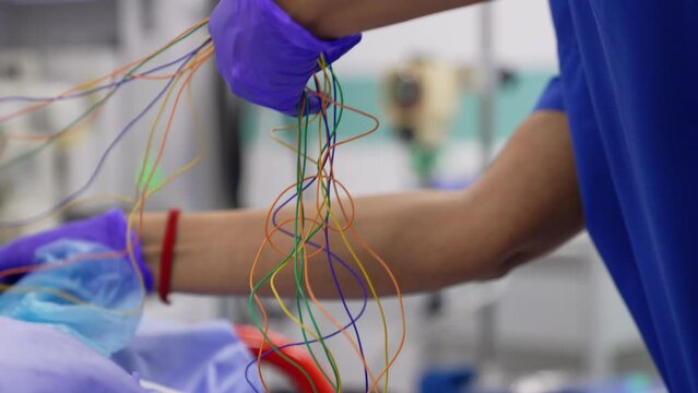 Gloved hands of doctor hold a few colorful thin wires. Medic attaches the equipment for neuromonitoring to a patient's head. Close up.