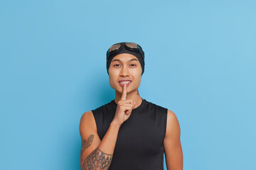 Asian swimmer in swimming cap, in black bathing top stands on blue background pressing index finger to his lips, sport life concept, copy space
