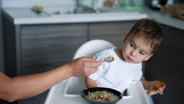 Mother is trying to feed her baby boy with soup. Kid looks attentively ahead and avoids spoon.