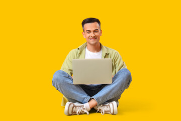 Happy young man sitting on floor with laptop against yellow background