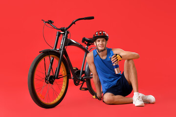 Young man sitting near bicycle and drinking water on red background