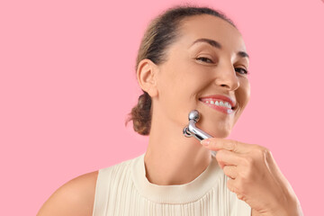 Beautiful mature woman using face massage roller on pink background