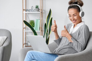 Mature woman with laptop and headphones showing thumb-up at home - obrazy, fototapety, plakaty