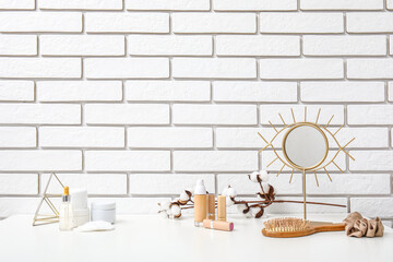 Different makeup products, hair comb and mirror  on dressing table near light brick wall in room