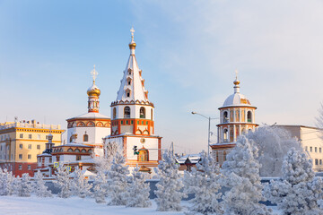  Irkutsk on frosty winter day. View from Lower Embankment of Angara River to beautiful Epiphany Cathedral in Old Siberian Baroque style. Snow-covered pine trees at pedestrian street. Winter cityscape