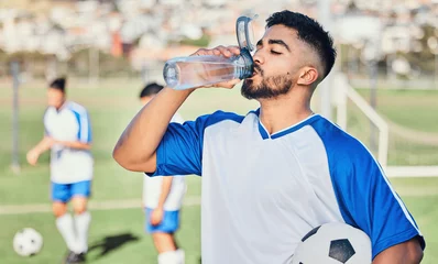 Fotobehang Football, athlete and man drinking water outdoor on a sports field for fitness competition. Tired male soccer player on a break and exhausted from exercise, challenge or training workout for health © C. Daniels/peopleimages.com
