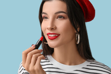 Beautiful young woman in beret applying red lipstick on blue background