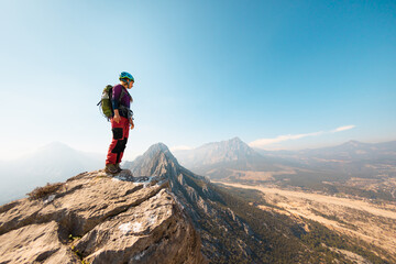 young girl climber in a helmet and with a backpack stands on top of a mountain. mountain climbing - obrazy, fototapety, plakaty