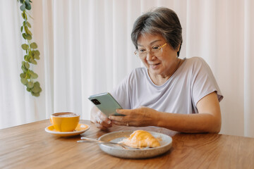 Happy asian old elder woman smiling and looking at mobile, holding and using phone, chatting or reading news, sitting on chair alone over white curtain at cafe with coffee and croissant on wood table.