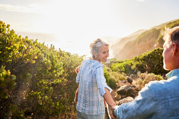 Naklejka premium Love, holding hands and nature, senior couple walking with picnic blanket for romance and valentines day. Green mountain path, old man and woman on walk or hike for romantic valentine date at sunset.