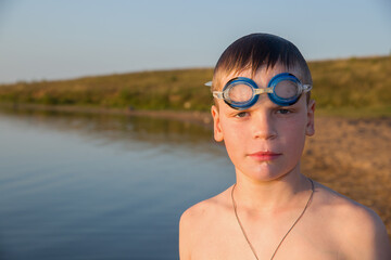 Portrait of a European boy wearing swimming goggles. In the background is the coastline of the sea.