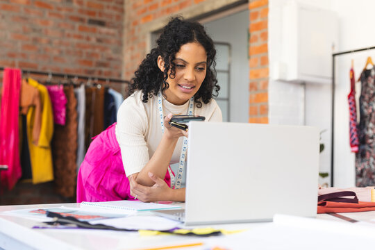 Happy biracial female fashion designer using smartphone and laptop in sunny studio - Powered by Adobe