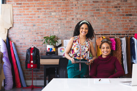 Two happy biracial female fashion designers using laptop, tablet and smiling in sunny studio