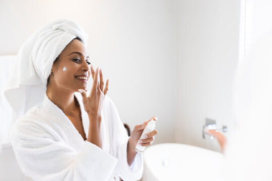 Happy biracial woman wearing bathrobe and applying cream in front of mirror in bathroom