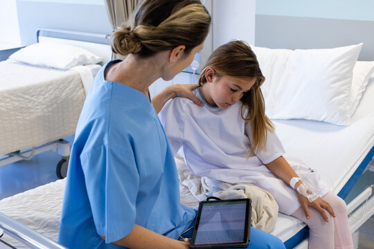 Caucasian female doctor with tablet comforting sad girl patient sitting on hospital bed
