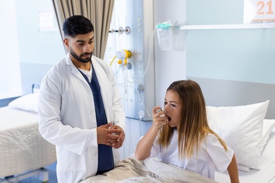 Diverse male doctor watching girl patient using asthma inhaler sitting up in hospital bed