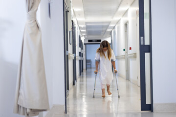Caucasian girl patient with head down walking with crutches in hospital corridor, copy space