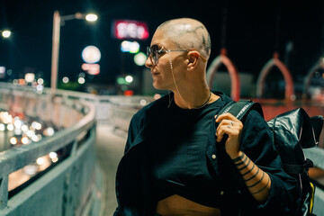 A serious young girl with no hair in a black dress and sunglasses, looks away