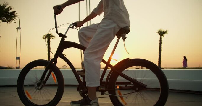 A happy man in white trousers pedals and rides his black bicycle along the morning beach along the sea at sunrise in summer