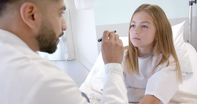 Diverse male doctor checking eyes of girl patient in hospital bed with penlight, slow motion