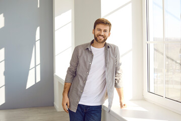 Portrait of a happy man at home. Smiling young man in a casual gray shirt, white T shirt and jeans standing by the window in a light clean room in his house or apartment