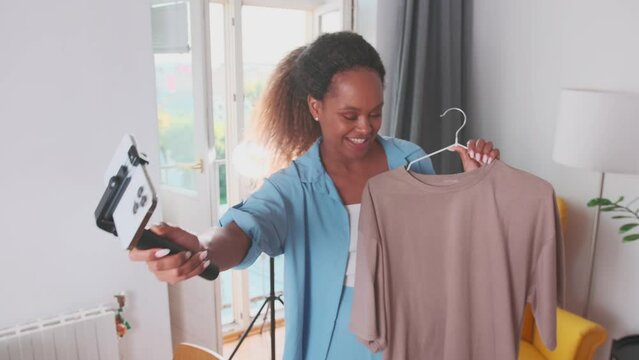 Young beautiful cheerful African American woman blogger records video broadcast on mobile phone telling subscribers about new t-shirt purchased in fashion boutique stands in apartment.