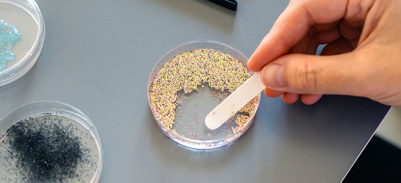 Banner of female scientist hands taking sample of small plastic particles from golden glitter on petri dish in environment research laboratory. Concept of analysis in microplastics composition.