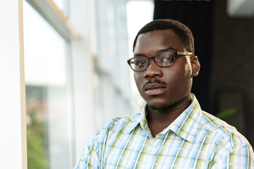 Portrait of thoughtful young african man in glasses standing near the window