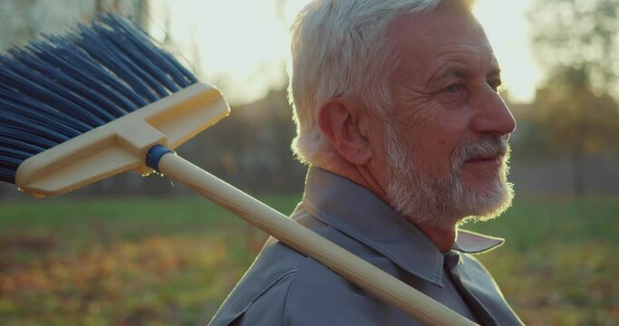 Senior caucasian concierge posing at city park with broom on shoulder. Grey haired man taking care of urban territory during autumn season.