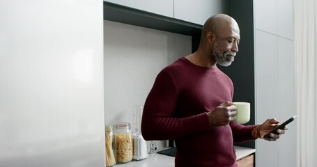 Happy biracial man drinking coffee and using smartphone in kitchen, slow motion - Powered by Adobe