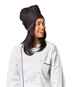 Young Filipina chef with cooking hat in studio looks aside smiling, cheerful and pleasant.