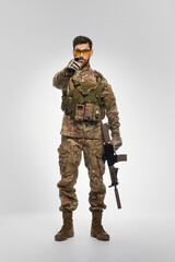 Severe male soldier with rifle pointing to camera with forefinger indoors. Front view of force officer in camouflage outfit, holding shotgun and warning, on gray backdrop. Concept of military force.